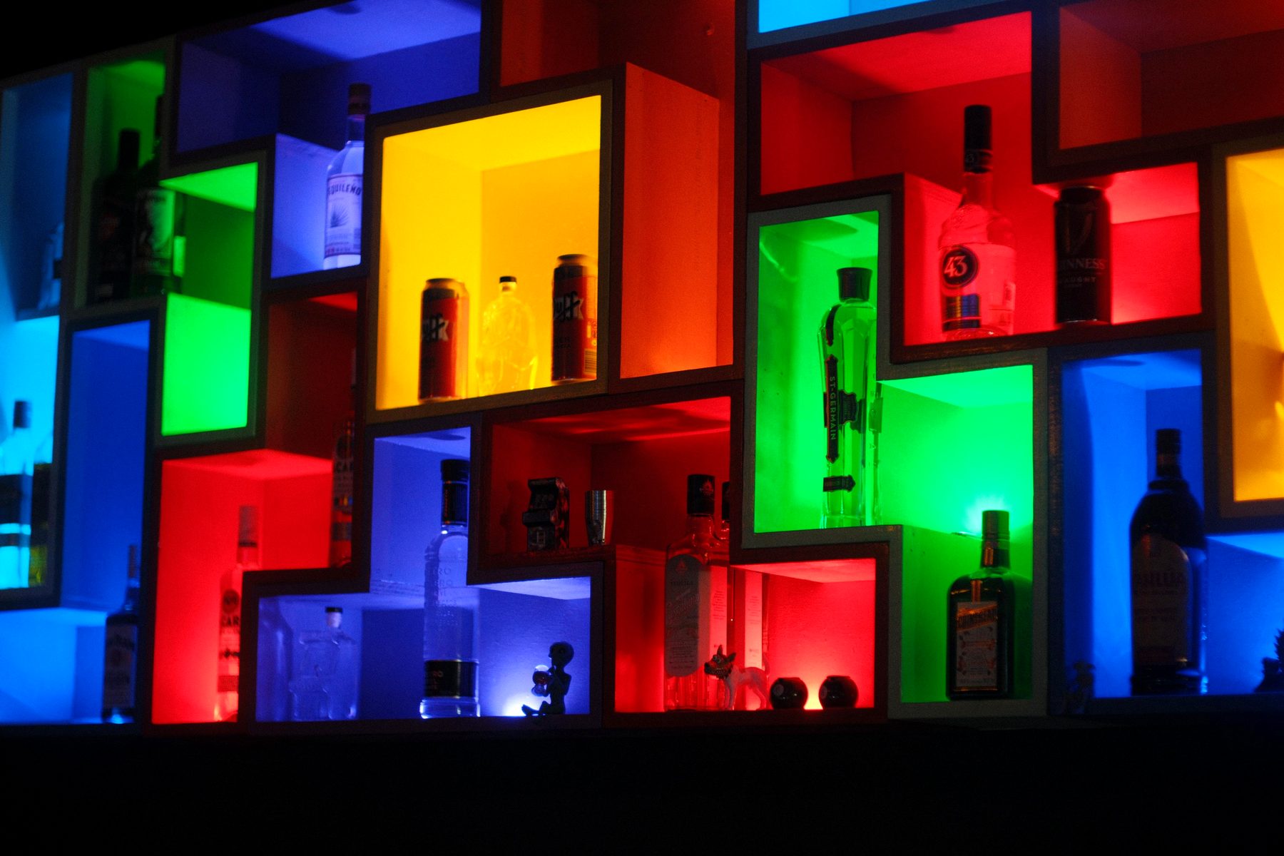 Pared de bloques con nichos iluminados en azul, verde, rojo, amarillo y naranja, con botellas dentro de cada pieza